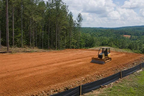 Small dozer leveling a homesite pad of packed red clay on a gentle slope with grade stakes set and a silt fence along the downhill edge