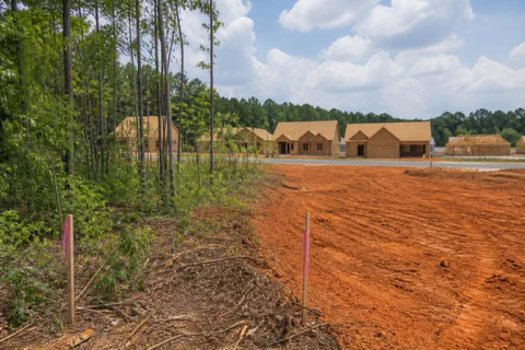 Half-cleared subdivision lot in Cabarrus County with graded red clay meeting standing pines, pink-flagged survey markers along the lot line
